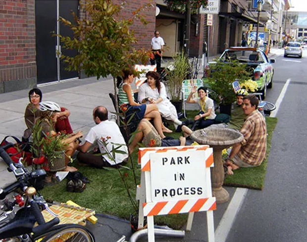 A group of people sit in a repurposed curbside parking space that they've outfitted with fake grass and trees.