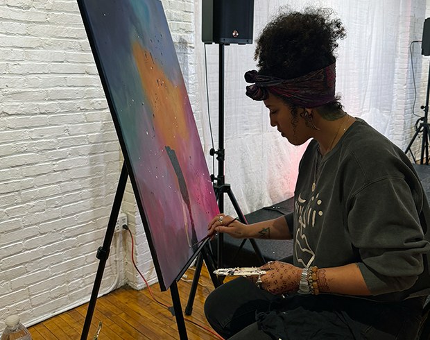 A woman with dark skin and hair pulled up leans over an easel while painting.