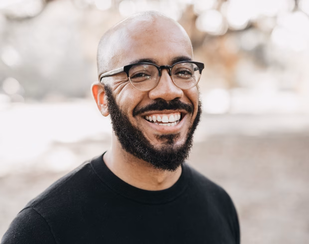 A young African American man with dark skin, a dark beard, and wearing glasses smiles at the camera.